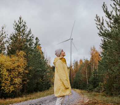 Nainen seisoo hiekkatiellä metsässä, taustalla tuulivoimala